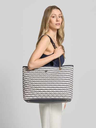 Frauenmodel trägt Tote Bag mit Viskose-Anteil Modell 'GIULLY' von Guess, blau-weiß gemustert, Logo-Detail, Nahaufnahme Oberkörper.