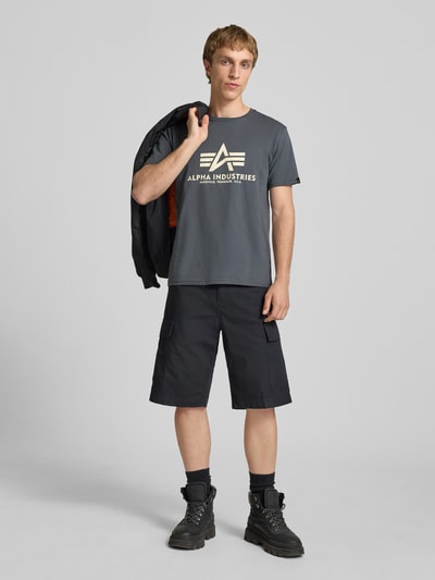 Männermodel trägt T-Shirt mit Logo und Rundhalsausschnitt von Alpha Industries in Dunkelgrau, Regular Fit, aus Baumwolle, Ganzkörperansicht von vorne.