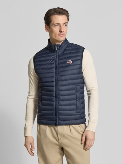 Męski model nosi Daunenweste mit Zweiwege-Reißverschluss od Colmar Originals, wypełnienie puchem i pierzem, krój regular fit, stójka, detal z logo, długość tyłu 62 cm, szerokość ramion 43 cm, szerokość w rozmiarze 50 klatki piersiowej 54 cm, granatowa, tekstura pikowana, widok całej sylwetki z przodu