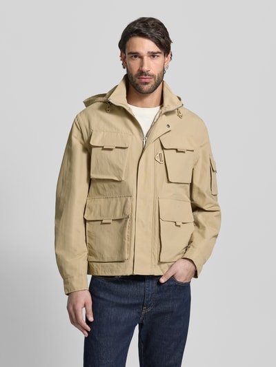 Männermodel trägt Oversized Funktionsjacke mit abnehmbarer Kapuze Modell 'BALTERO2611' von HUGO in Beige, unifarben, wasserabweisend, mehrere Taschen, Langarm mit Manschette, Ganzkörperansicht von vorne.