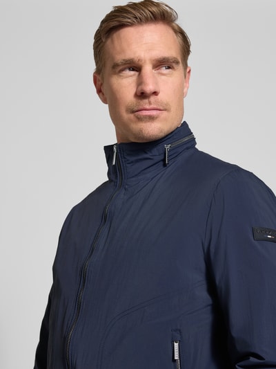Męski model nosi Regular Fit Jacke mit Stehkragen od Bugatti, granatowy kolor, zamek błyskawiczny dwustronny, detal z logo, kaptur, wpuszczane kieszenie zapinane na zamek błyskawiczny, zakończenia rękawów elastyczne, długość tyłu w rozmiarze 50: 65 cm, szerokość ramion w rozmiarze 50: 47 cm, szerokość klatki piersiowej w rozmiarze 50: 53 cm, długość rękawów w rozmiarze 50: 63 cm, widok z przodu, zbliżenie na górną część ciała.