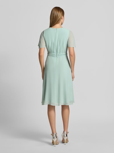 Frauenmodel trägt Knielanges Cocktailkleid in Wickel-Optik von Christian Berg Cocktail aus mintfarbenem Chiffon, A-Linie, Bindegürtel, Ganzkörperansicht Rückansicht.