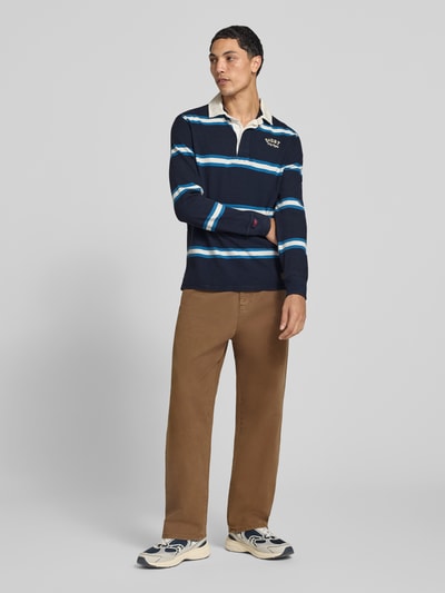 Männermodel trägt Poloshirt aus reiner Baumwolle von Superdry in Dunkelblau mit hellblauen Streifen, gerader Schnitt, Ganzkörperansicht von vorne. Rückenlänge bei Größe M: 71 cm, elastische Abschlüsse, Label-Stitching, Polokragen mit Abdeckleiste.