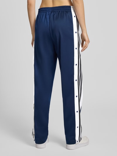 Frauenmodel trägt Straight Fit Jogginghose mit elastischem Bund Modell 'Adibreak' von adidas Originals in Dunkelblau mit weißen Streifen, Ziernähte und Eingrifftaschen, Ganzkörperansicht Rückansicht.