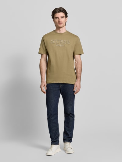 Männermodel trägt T-Shirt mit Label-Detail von Guess aus reiner Baumwolle in Oliv, Regular Fit, gerippter Rundhalsausschnitt, Ganzkörperansicht von vorne.