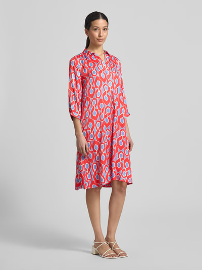 Frauenmodel trägt Knielanges Hemdblusenkleid mit V-Ausschnitt von Milano Italy aus reiner Viskose in Rot-Blau mit grafischem Allover-Muster, A-Linie, Hemdblusenkragen, knielang, Ganzkörperansicht von vorne.