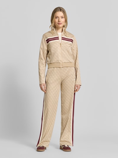 Frauenmodel trägt Regular Fit Sweatjacke mit Allover-Logo-Print Modell 'Jacquard' von Guess, beige mit braunen Streifen, Baumwoll-Mix, Stehkragen, Reißverschluss, elastischer Bund, Ganzkörperansicht von vorne.