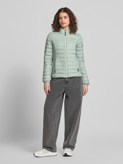 Frauenmodel trägt mintgrüne Steppjacke mit Logo-Stitching Modell 'FUJI' von Superdry, Gerader-Schnitt, Stehkragen, Reißverschluss, Seitliche Reißverschlusstaschen, Rückenlänge 62 cm, Schulterbreite 40 cm, Brustweite 50 cm, Ärmellänge 69 cm, Ganzkörperansicht von vorne.