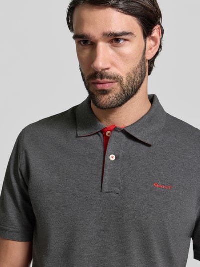 Męski model nosi Regular Fit Poloshirt mit Logo-Stitching od Gant, elastyczna bawełna, szary kolor, krótkie rękawy, haftowane logo, krój regular fit, zbliżenie na górną część ciała.