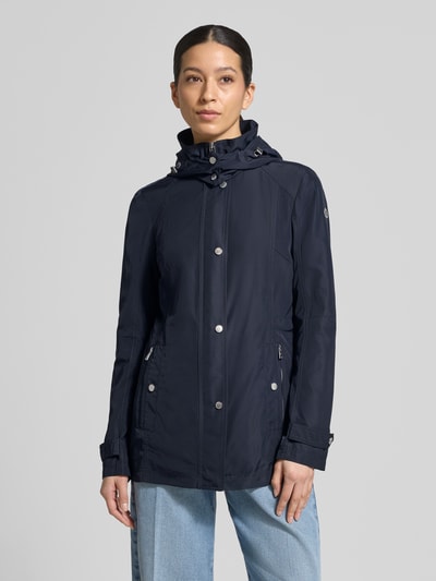 Frauenmodel trägt Regular Fit Jacke mit Logo-Print von Fuchs Schmitt in Dunkelblau, glatte Textur, silberne Reißverschlüsse, Ganzkörperansicht von vorne. Rückenlänge 71 cm, Schulterbreite 40 cm, Brustweite 51 cm, Ärmellänge 66 cm, Stehkragen, Kapuze mit Tunnelzug, Eingrifftaschen.
