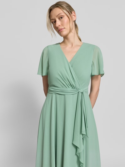 Vrouwenmodel draagt knielange cocktailjurk met strikceintuur van Christian Berg Cocktail, chiffon, getailleerd model, wikkellook, nahaufnahme bovenlichaam.