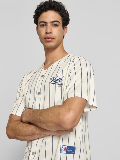 Männermodel trägt Regular Fit Freizeithemd mit Logo-Stitching von KARL KANI in Creme mit dunkelblauen Streifen, Kurzärmelig, Nahaufnahme Oberkörper. Rückenlänge 64 cm, Schulterbreite 45 cm, Ärmellänge 27 cm, V-Ausschnitt, durchgehende Knopfleiste.