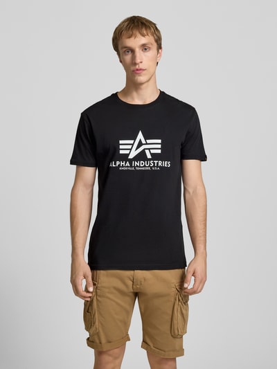 Männermodel trägt schwarzes T-Shirt mit Logo und Rundhalsausschnitt von Alpha Industries aus Baumwolle, Regular Fit, Ganzkörperansicht von vorne.