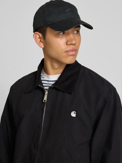 Männermodel trägt Jacke mit Label-Stitching Modell 'Madison' von Carhartt Work In Progress aus schwarzer Baumwolle, Nahaufnahme Oberkörper. Regular Fit, Reißverschluss, dezente Streifen, glatte Textur, Eingrifftaschen, neutrale Farbtöne, Kontextdaten integriert.