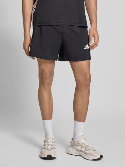 Männermodel trägt Shorts mit Logo-Print und Reißverschlusstaschen von adidas Training, schwarz mit weißem Logo, Lockerer-Schnitt, Elastikbund, Seitliche Reißverschlusstaschen, Bundweite 38 cm, Innenbeinlänge 14 cm, Ganzkörperansicht von vorne.