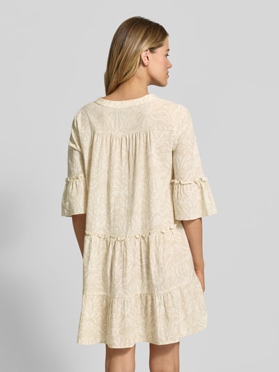 Vrouwenmodel draagt ausgestelltes Blusenkleid aus reiner Baumwolle Modell 'DICTHE' van Vero Moda met allover-patroon, mini lengte, ruches, Henley-halslijn en trompetmouwen, lichte beige tinten, zachte katoenen textuur, uitlopende fit. Volledige lichaamsweergave achterkant.