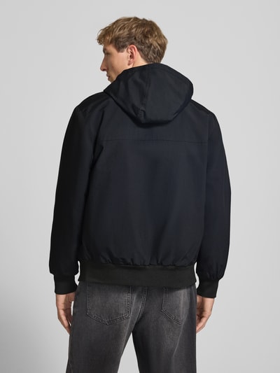 Männermodel trägt schwarze Funktionsjacke mit Logo-Stitching und Kapuze von Carhartt Work In Progress, gerader Schnitt, Ganzkörperansicht Rückansicht.