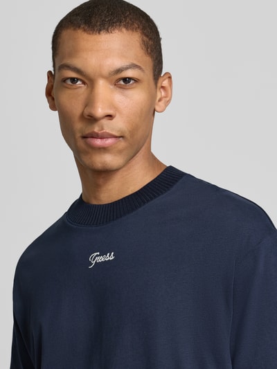 Männermodel trägt T-Shirt mit Label-Detail von Guess in Dunkelblau aus reiner Baumwolle mit geripptem Rundhalsausschnitt, Nahaufnahme Oberkörper. Lockerer Schnitt, Rückenlänge bei Größe M 73 cm.