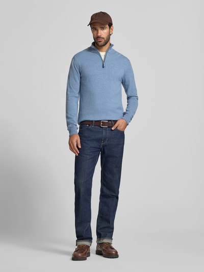 Männermodel trägt Pullover aus reiner Wolle von GANT in Hellblau, Troyer-Kragen mit Reißverschluss, Feinstrick, lange Ärmel, Ganzkörperansicht von vorne.