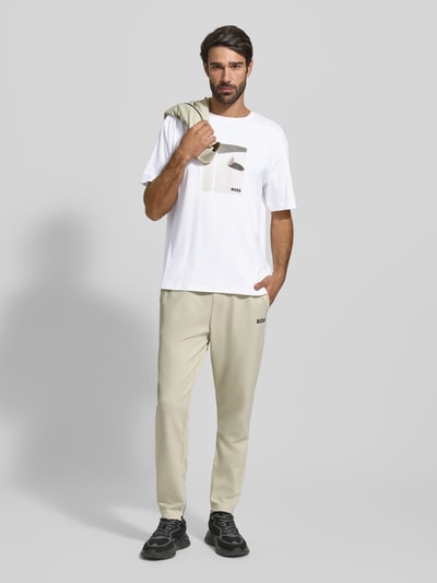 Męski model nosi Relaxed Fit T-Shirt aus reiner Baumwolle Modell 'JOIN' od BOSS Green, biała koszulka z nadrukiem i haftem, widok całej sylwetki z przodu.
