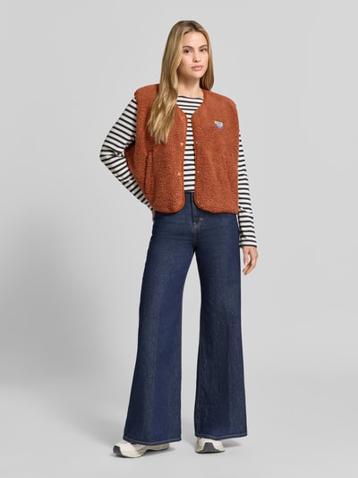 Frauenmodel trägt Relaxed Fit Weste aus Teddyfell Modell 'Hoktown' von American Vintage in Braun, weiße Knopfleiste, Logo-Patch; dazu gestreiftes Langarmshirt und blaue Jeans, Outfit farblich kontrastreich, strukturierte Oberfläche, Ganzkörperansicht von vorne. Damen Weste von American Vintage, Relaxed Fit, Rückenlänge 56 cm, Schulterbreite 43 cm, Brustweite 63 cm.