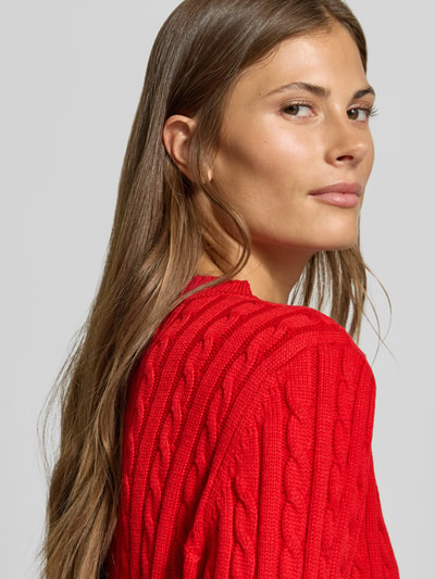 Frauenmodel trägt Strickjacke mit Woll-Anteil von MYON in leuchtendem Rot mit Zopfmuster, gerippte Abschlüsse, Nahaufnahme Oberkörper.