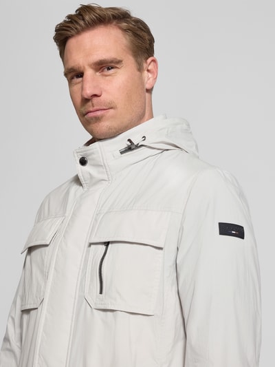 Mężczyznamodel nosi Regular Fit Jacke mit Stehkragen od Bugatti w jasnoszarym kolorze, z kieszeniami na piersi, chowanym kapturem, aplikacją z logo; zbliżenie na górę sylwetki. Długość tyłu w rozmiarze 50: 72 cm, szerokość ramion w rozmiarze 50: 46 cm, szerokość w rozmiarze 50: 52 cm, długość rękawów w rozmiarze 50: 68 cm.