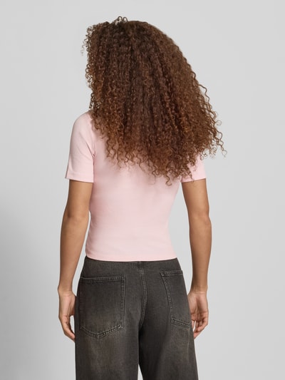 Modelka nosi jasnoróżowy Slim Fit T-Shirt aus Baumwoll-Mix Modell 'FEMKE' od Only, gładki, krój slim fit, krótkie rękawy, dekolt okrągły, drobne detale w formie kokardek, dzianina prążkowana, materiał bawełniany z domieszką, widok całej sylwetki od tyłu.