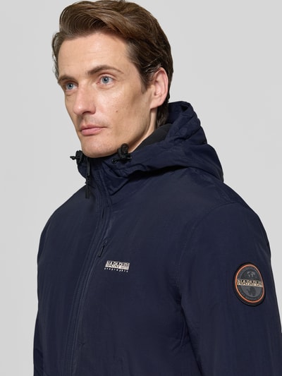 Männermodel trägt Jacke mit Label-Print Modell 'TUNDRA' von Napapijri in Dunkelblau, sichtbarer Logo-Patch, Nahaufnahme Oberkörper. Gerade Passform, Kapuze mit Tunnelzug, durchgehender Reißverschluss, Reißverschlusstaschen, elastische Ärmelabschlüsse, dezente strukturierte Oberfläche.
