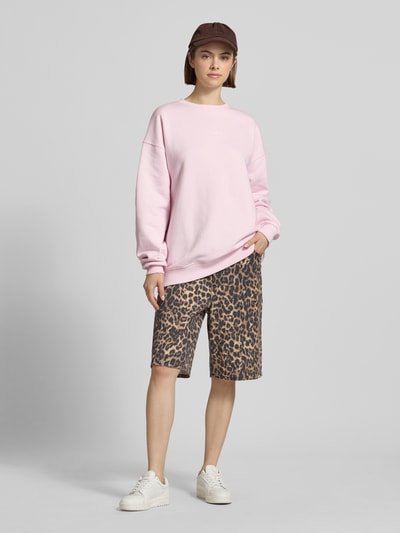 Frauenmodel trägt Oversized Sweatshirt mit Logo-Print von OH APRIL aus Baumwoll-Mix, Oversized-Fit, gerippter Rundhalsausschnitt, überschnittene Schultern, hellrosa Farbe, glatte Textur, kombiniert mit gemusterten Shorts, Ganzkörperansicht von vorne.
