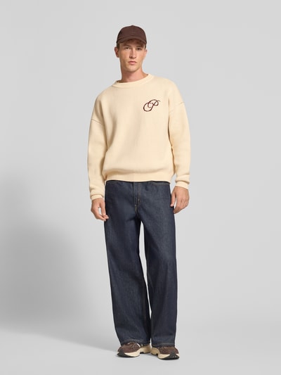 Männermodel trägt Relaxed Fit Strickpullover aus reiner Baumwolle von PROHIBITED in Creme mit Label-Stitching, Ganzkörperansicht von vorne.