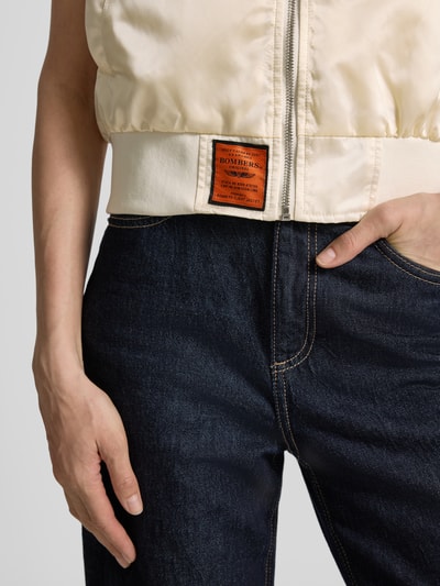 Frauenmodel trägt Weste mit Reißverschluss und Label-Detail von Original Bombers in Creme, sichtbar sind Stehkragen und Eingrifftaschen, Nahaufnahme Oberkörper.