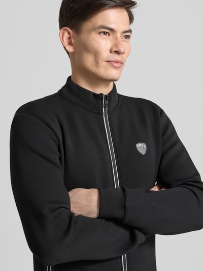 Männermodel trägt Sweatjacke mit Reißverschluss von EA7 Emporio Armani, Regular-Fit, gerippte Abschlüsse, Stehkragen, Label-Detail, Eingrifftaschen, schwarze Farbe, Nahaufnahme Oberkörper.