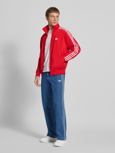 Männermodel trägt Sweatjacke mit Logo-Stitching Modell 'FIREBIRD' von adidas Originals in Rot mit weißen Details, Ganzkörperansicht von vorne. Herren Sweatjacke, Gerader-Schnitt, Stehkragen, Reißverschluss, seitliche Reißverschlusstaschen, elastische Abschlüsse, Rückenlänge Größe M: 72 cm, Ärmellänge: 67 cm.