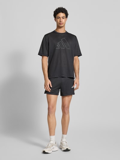 Męski model nosi Straight Fit Shorts mit Logo-Print in Mesh-Optik von adidas Training, czarne, krój straight fit, szerokość w rozmiarze M: 39 cm, długość wewnętrzna nogawki: 12 cm, widok całej sylwetki z przodu.