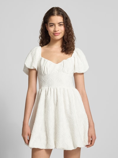 Vrouwenmodel draagt Minikleid met floralem Loch-Stitching van Gina Tricot in wit, elastische materiaalmix, getailleerd, plooien-look, jurk met korte mouwen. Ganzkörperansicht van voren.