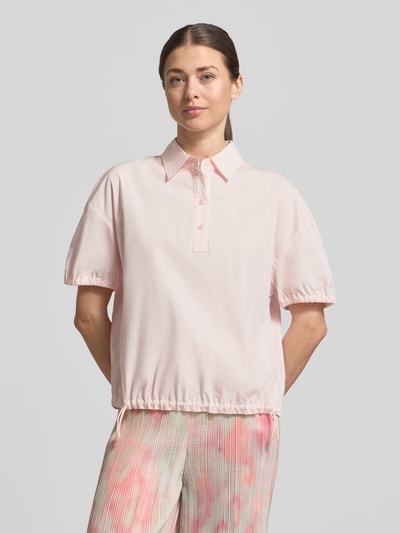 Modelka kobieca nosi Regular Fit Bluse mit Polokragen od Marc Cain z bawełny, krój regular fit, kołnierzyk polo, krótkie rękawy, elastyczne zakończenia rękawów, pas z tunelem, pastelowy róż, długość tyłu w rozmiarze 36: 59 cm, długość rękawa: 19 cm, widok całej sylwetki z przodu.