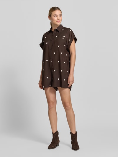 Frauenmodel trägt Lockeren Jumpsuit mit Knopfdetail – curated by Mary Braun von Style Icon Collection im Braun mit Allover-Look, Hemdblusenkragen, Knopfleiste und Relaxed-Fit, Ganzkörperansicht von vorne. Rückenlänge bei Größe S: 65 cm, Brustweite 60 cm, Ärmellänge 14 cm, Gesamtlänge 84 cm, Innenbeinlänge 5 cm.