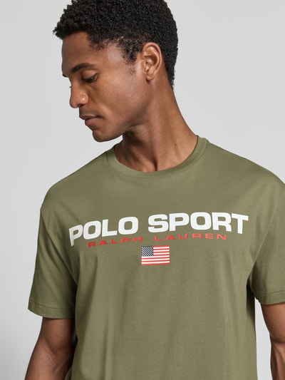 Męski model nosi Regular Fit T-Shirt aus reiner Baumwolle von Polo Ralph Lauren w kolorze oliwkowym, nadruk z flagą USA, zbliżenie na górną część ciała.