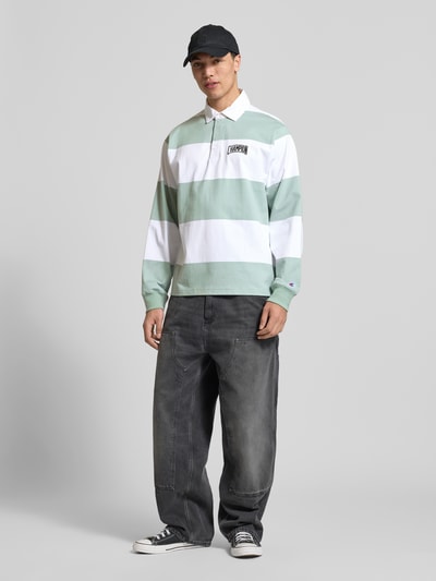 Männermodel trägt Relaxed Fit Poloshirt aus Baumwolle von CHAMPION, grün-weißes Streifendesign, Label-Stitching, Langärmlig, Ganzkörperansicht von vorne. Rückenlänge bei Größe M: 72 cm.