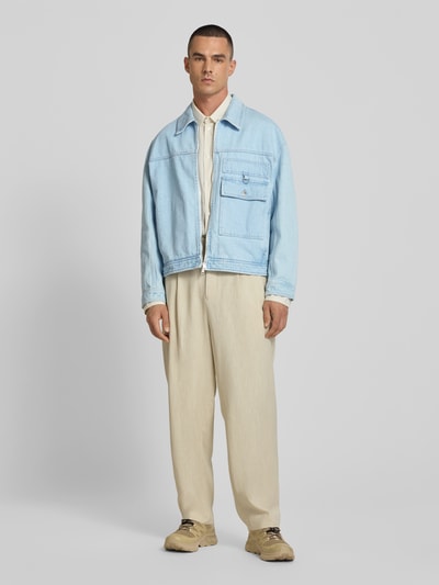 Männermodel trägt Jeansjacke aus reiner Baumwolle von MAISON KITSUNÉ in Hellblau mit Zweiwege-Reißverschluss, Gerade Passform, Brusttasche, Umlegekragen, beige Hose, neutrale Farben, Ganzkörperansicht von vorne.