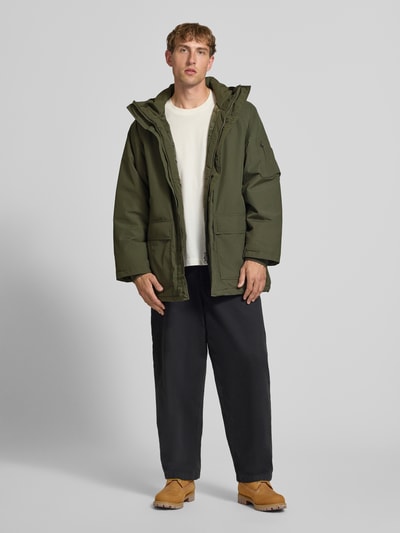 Männermodel trägt olivgrünen Parka mit Pattentaschen Modell 'Stanwood' von Carhartt Work In Progress, Ganzkörperansicht von vorne. Regular Fit mit Kapuze, Reiß- und Klettverschluss, Reißverschlusstasche am Ärmel und Label-Patch, glatte Textur.