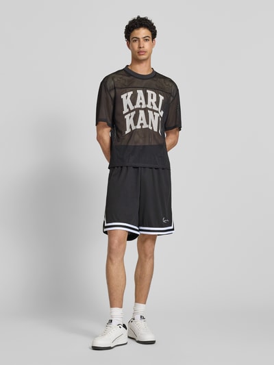 Männermodel trägt Relaxed Fit Shorts mit Logo-Stitching von KARL KANI aus Mesh mit Elastikbund in Schwarz-Weiß, Ganzkörperansicht von vorne.