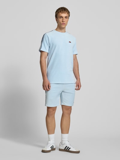 Męski model nosi T-Shirt mit Logo Modell 'MADORI' od Ellesse, mieszanka bawełny z dodatkiem streczu, krój regular fit, okrągły dekolt, logo, długość tyłu w rozmiarze M: 66 cm, jasnoniebieski kolor, prosty fason, tekstura prążkowana, całkowity widok z przodu.