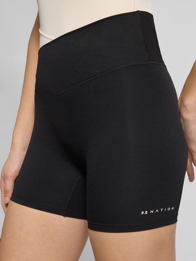 Frauenmodel trägt Slim Fit Shorts mit Logo-Print Modell 'Wander' von P.E Nation aus Elastischer-Material-Mix in Schwarz, Nahaufnahme Oberschenkel. Damen Shorts mit Elastischer-Logo-Bund, Logo-Print, Bundweite Größe S 29 cm, Innenbeinlänge 12 cm.