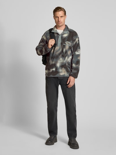 Männermodel trägt Fleecepullover mit Label-Patch Modell 'HELVETIA' von Columbia, gemustert in Grau- und Blautönen, Gerader Schnitt, Stehkragen, kurze Knopfleiste, Brusttasche, Jeans, Ganzkörperansicht von vorne.