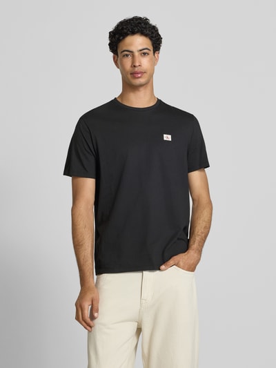 Männermodel trägt T-Shirt mit Label-Badge von Calvin Klein Jeans in Schwarz aus reiner Baumwolle, Regular Fit, Nahaufnahme Oberkörper.