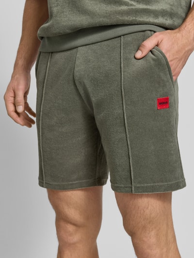 Männermodel trägt Regular Fit Sweatshorts aus Baumwoll-Mix Modell 'TERRANCE' von HUGO in Olivgrün mit Label Patch, Nahaufnahme Oberschenkelbereich.