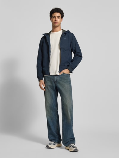Männermodel trägt Regular fit Jack met logoprint model 'Track' van G-Star Raw in donkerblauw met steekzakken en capuchon, ganzkörperansicht von vorne. Ruglengte maat M 65 cm, schouderbreedte 41 cm, borstwijdte 48 cm, mouwlengte 67 cm.
