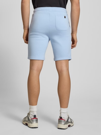 Mannenmodel draagt Shorts met Eingrifftaschen und Logo-Print van Alpha Industries in lichtblauw katoenmix, relaxed fit, achterzak, ganzkörperansicht rückansicht. Context: heren short met elastische band met trekkoord, zij-steekzakken, logoprint, bandwijdte maat M 39 cm, binnenbeenlengte 24 cm.